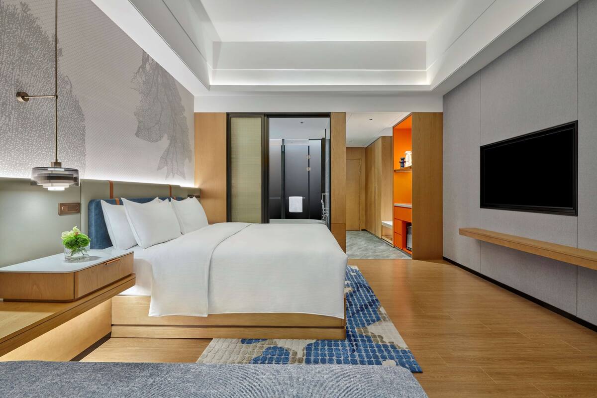A modern hotel room with a large bed, bedside tables, a TV, and a view of the hallway.