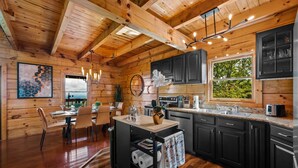A wooden cabin interior with a kitchen area featuring a dining table, chairs, a black cart with towels, and a large window with a view of greenery. - Ridgetop VIEWS! Arcade+Hot Tub+Fire Pit+Privacy (Sevierville)