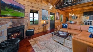 A cozy log cabin interior with a stone fireplace, a leather sofa, and a large flat-screen TV. - Ridgetop VIEWS! Arcade+Hot Tub+Fire Pit+Privacy (Sevierville)