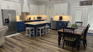 Una cocina moderna con armarios blancos, un refrigerador de acero inoxidable, una isla central con taburetes y un comedor con mesa y sillas de madera.