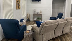 Una sala de estar con un sofá color crema, un sillón azul y una mesa de centro de madera. Hay un televisor montado en la pared, una mesita auxiliar con una lámpara y una ventana con persianas.