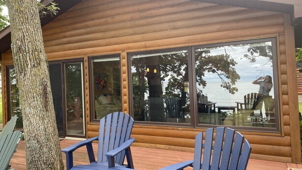 Una cabaña de madera con ventanas amplias, una terraza con dos sillas Adirondack y un árbol en primer plano.