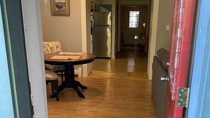 A hallway with a wooden floor leading to a dining area with a table and chairs. A refrigerator is visible in the background.