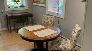 A dining area with a round wooden table, two chairs, a lamp, a map on the wall, and a window with blinds.