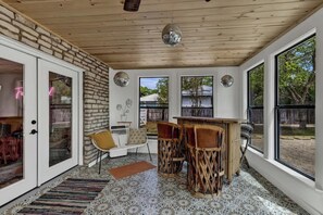 A cozy room with a stone wall, a wooden ceiling, and large windows. There is a white sofa, a small table, and two chairs.