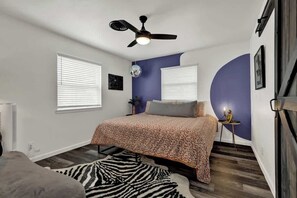 Ein Schlafzimmer mit Bett, Deckenventilator, zwei Fenstern mit Jalousien, einem zebra-gemusterten Teppich und einem kleinen Tisch mit einer Statue.