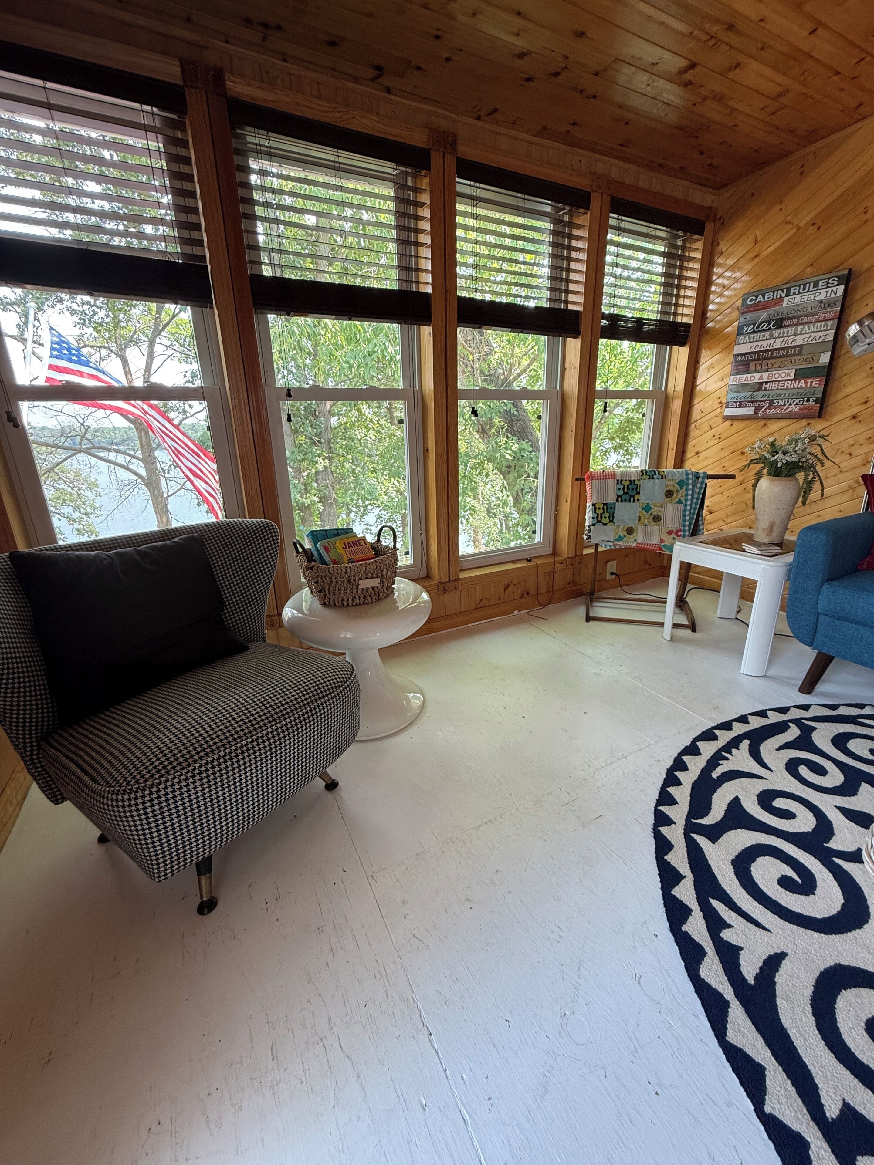Interior de una cabaña con un sofá a cuadros de piedritas, una mesa de centro blanca, un sillón azul y un ventanal con persianas.