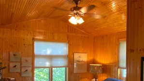 Un dormitorio de cabaña de madera con una cama, un ventilador de techo y una ventana con persianas.