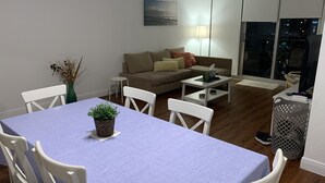 Ein modernes Wohnzimmer mit Esstisch, weißen Stühlen, einer Couch und einem Couchtisch. Auf dem Esstisch steht eine Pflanze, auf der Couch eine Lampe, und durch das Fenster hat man Blick auf die beleuchtete Stadt.