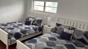 Ein Schlafzimmer mit zwei Betten, jeweils mit einem Bettbezug mit geometrischem Muster und passenden Kissen. An einer Seite befindet sich ein Nachttisch mit einer Lampe und eine kleine Schublade. Über den Betten ist ein Fenster mit Jalousien zu sehen.