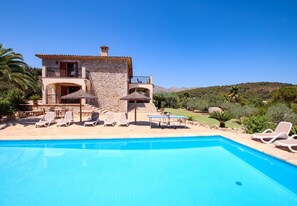 A poolside view of a stone building with a balcony, surrounded by lounge chairs and a clear blue sky.