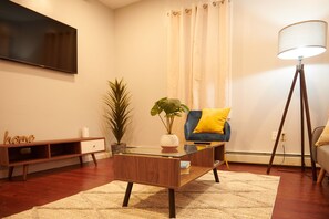 A modern living room with a glass coffee table, a blue sofa with a yellow pillow, a floor lamp, and a flat-screen TV mounted on the wall. - Cozy apartment 2nd 10min Walk Downtown | City View (Providence)