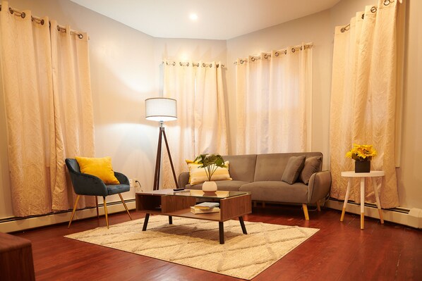 A living room with a sofa, a coffee table, a chair, a lamp, and a small table with a vase of flowers. - Cozy apartment 2nd 10min Walk Downtown | City View (Providence)