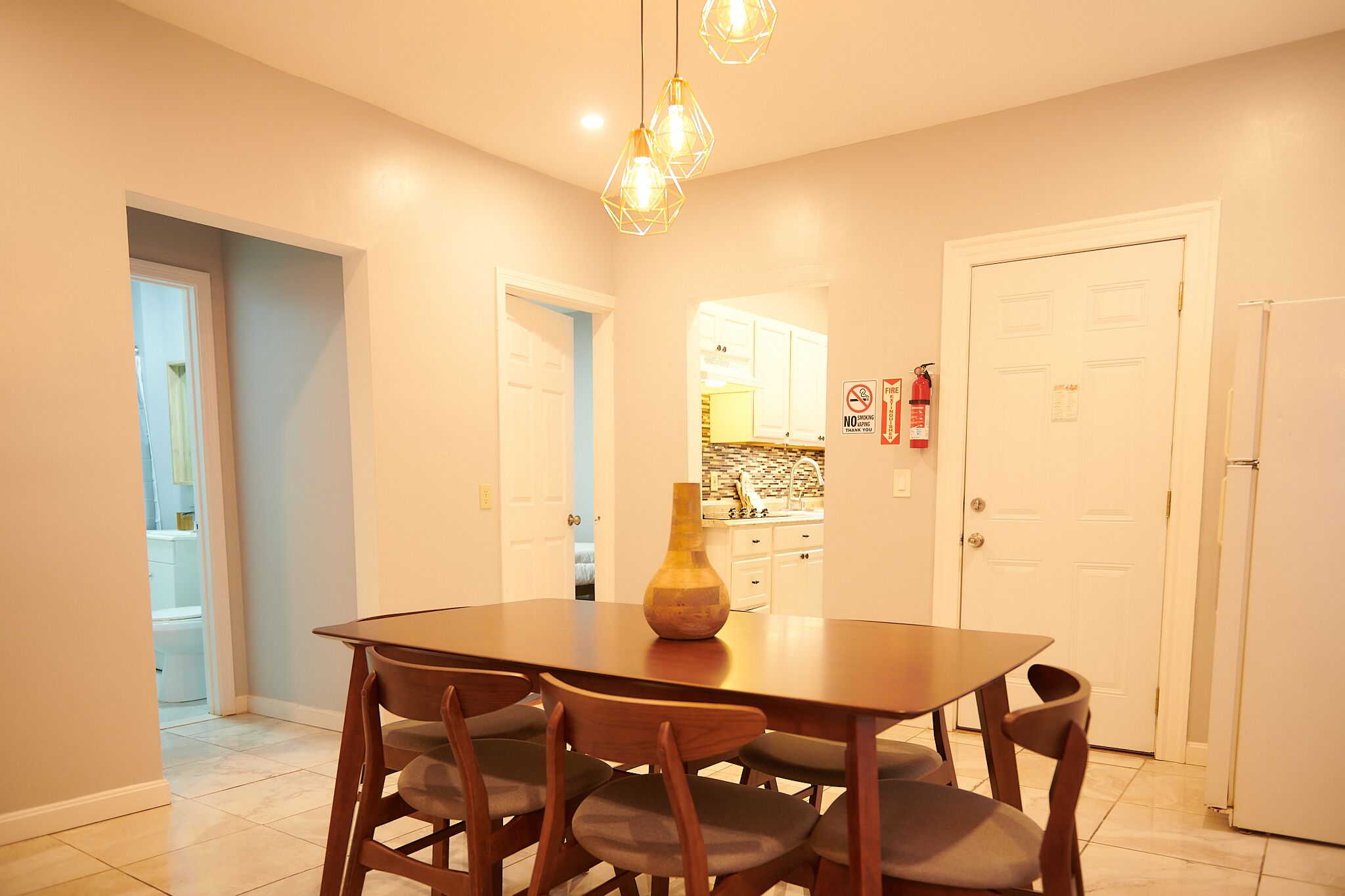 A dining area with a wooden table and chairs, a vase on the table, and a kitchen in the background.