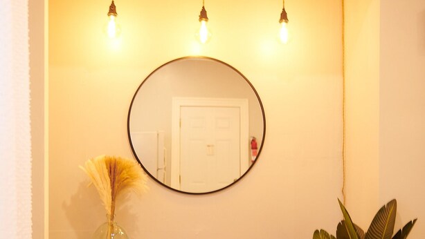 A wooden console table with a mirror above it, flanked by two hanging light bulbs and a potted plant to the right.