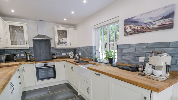 A modern kitchen with white cabinets, a built-in oven, a coffee machine, and a window with a view of greenery. - Carreg Winllan (Pen-y-sarn)
