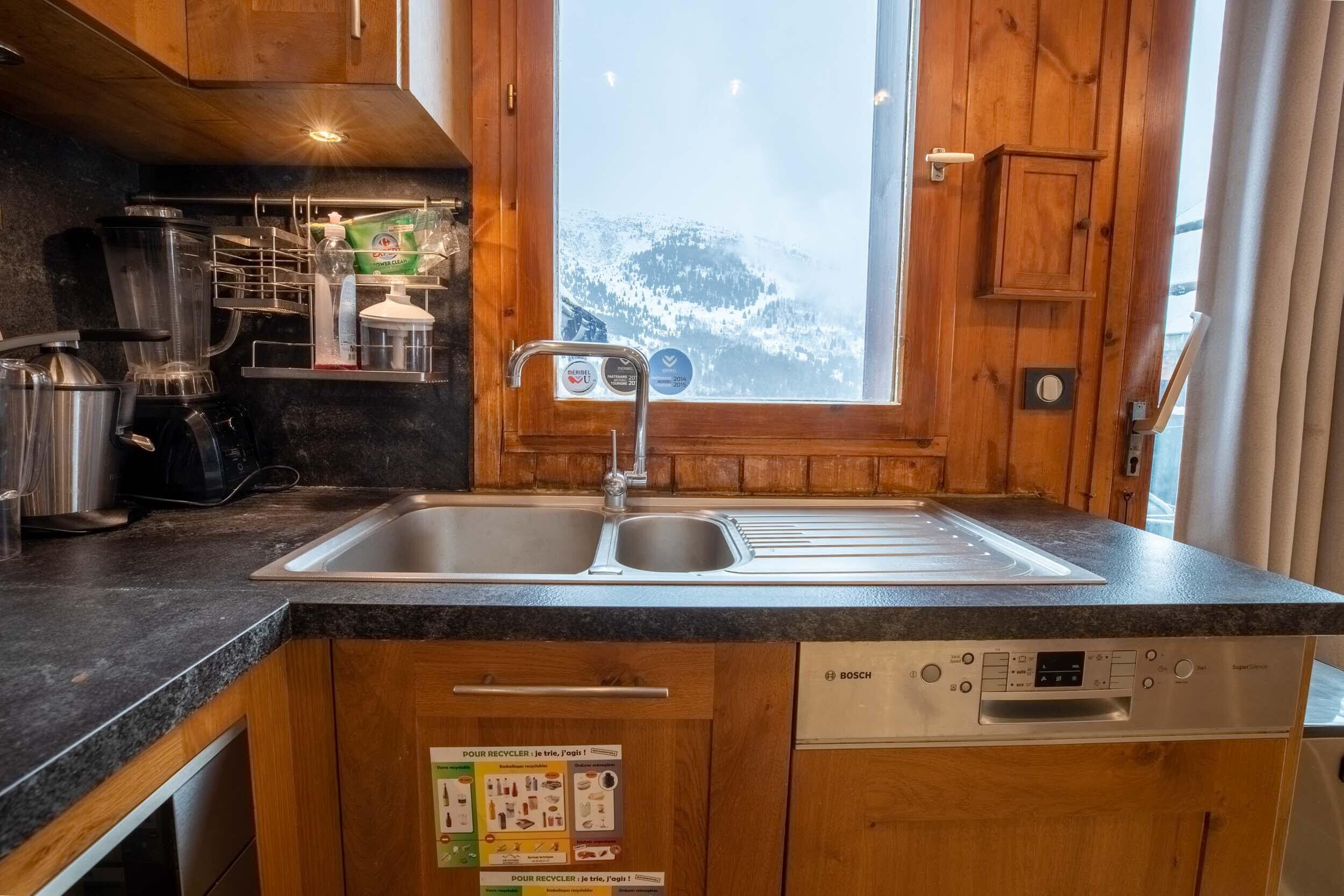 Een keuken met houten kastjes, een Bosch vaatwasser en een raam met uitzicht op een besneeuwd berglandschap.