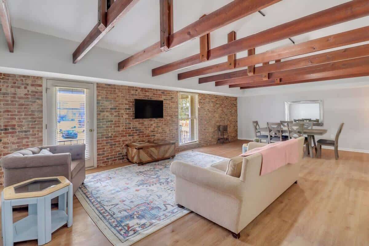 Ein geräumiges Wohnzimmer mit freiliegenden Holzbalken, einer Ziegelwand und einem großen Fenster. Es gibt ein beiges Sofa, einen kleinen Tisch und einen gemusterten Teppich.