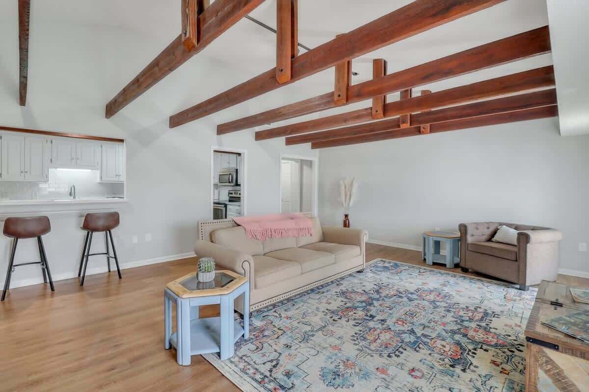 A spacious living area with exposed wooden beams, a beige sofa, a patterned rug, and a kitchenette in the background.
