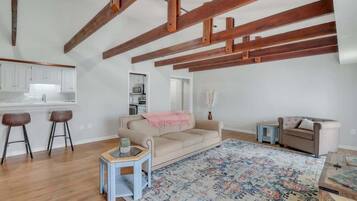 A spacious living area with exposed wooden beams, a beige sofa, a patterned rug, and a kitchenette in the background.