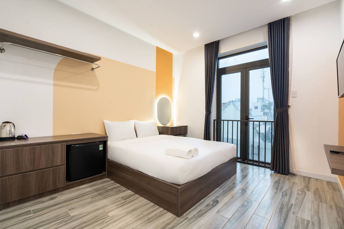 A modern hotel room with a large bed, a built-in desk, a small refrigerator, and a balcony with curtains.