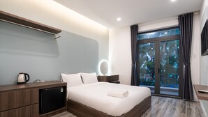 A modern hotel room with a wooden bed, a small refrigerator, a bedside table with a lamp, and a large window with curtains. - Justay Pink Church (Ho Chi Minh City)