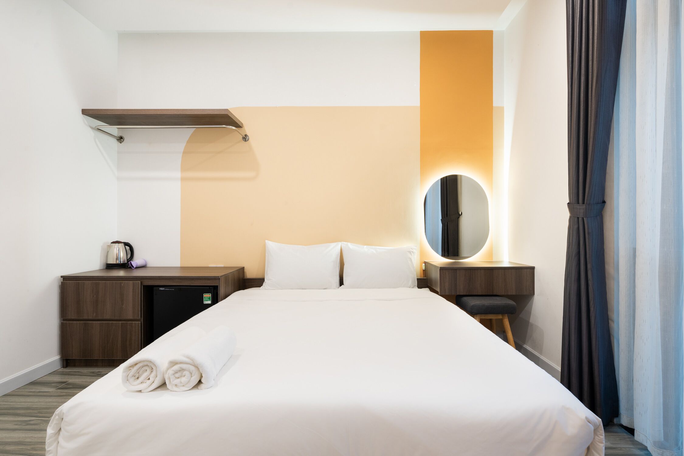 A hotel room with a neatly made bed, a small desk with a kettle, a mirror, and a nightstand.