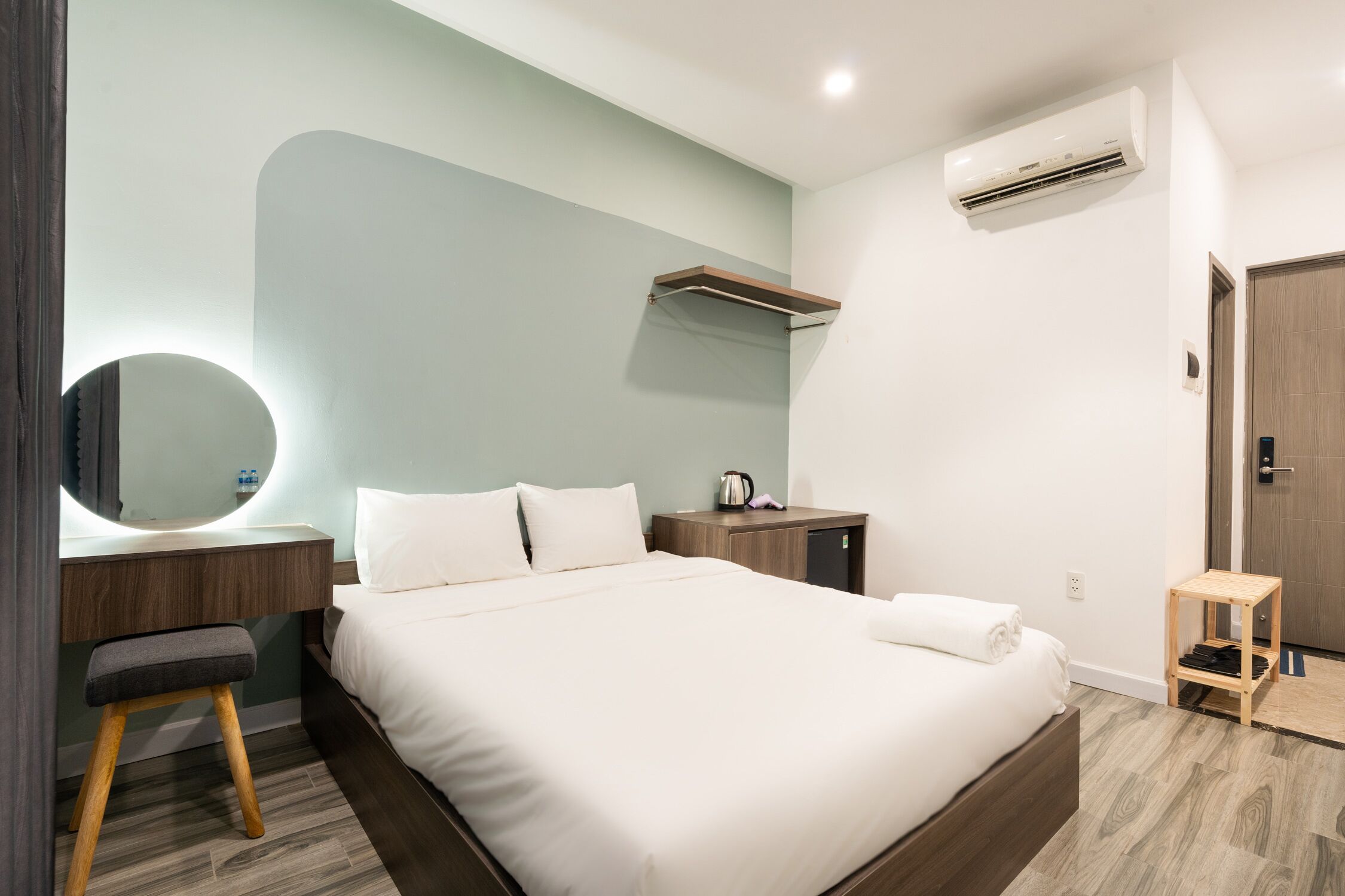 A modern hotel room with a large bed, a bedside table with a mirror, a small wooden stool, a nightstand, and a built-in shelf.