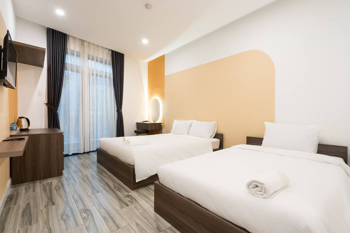 A hotel room with two beds, a desk, a vanity with a mirror, and a window with curtains.