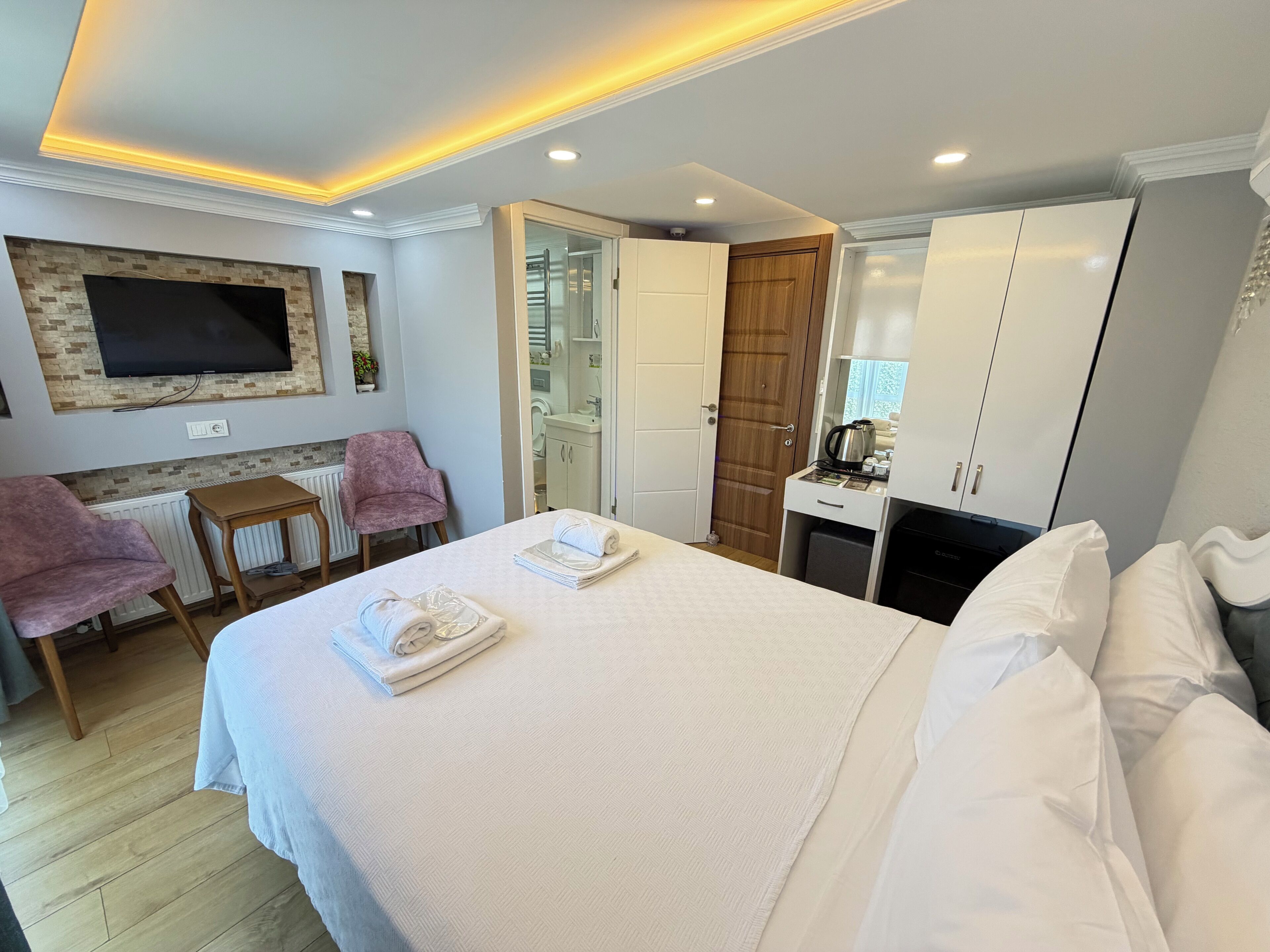 Habitación de hotel con una cama, un televisor montado en la pared, una puerta de madera, una mesita pequeña y una zona de estar con dos sillas.