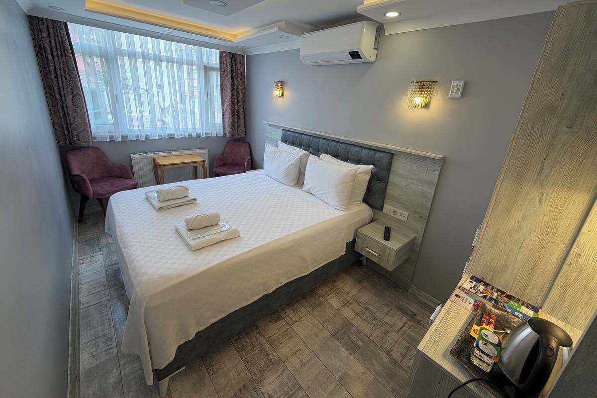 A hotel room with a bed, a chair, a table, an air conditioner, and a kettle.