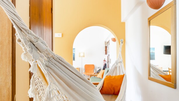 A hammock chair in a room with a wooden door, a mirror, and a view of a living area with a sofa and orange cushions. - Altura Inn Beach - Sol, na Praia, Varanda, Free Wifi e Ar Condicionado (Altura)