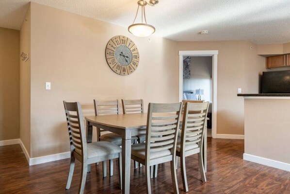 Un comedor con una mesa redonda y seis sillas, un reloj de pared y una zona de cocina al fondo.