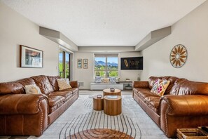 A spacious living room with leather sofas, a large window with a mountain view, a flat-screen TV, and a decorative wall piece.