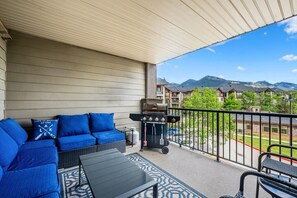 A balcony with a sofa, grill, and a view of buildings and mountains. - Poolside Pad | Pool/Hot Tub | Mountain Views | Gym (Invermere)