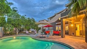 Piscina com espreguiçadeiras e guarda-sóis, rodeada por um espaço de terraço com mobiliário para área externa e uma casa ao fundo.