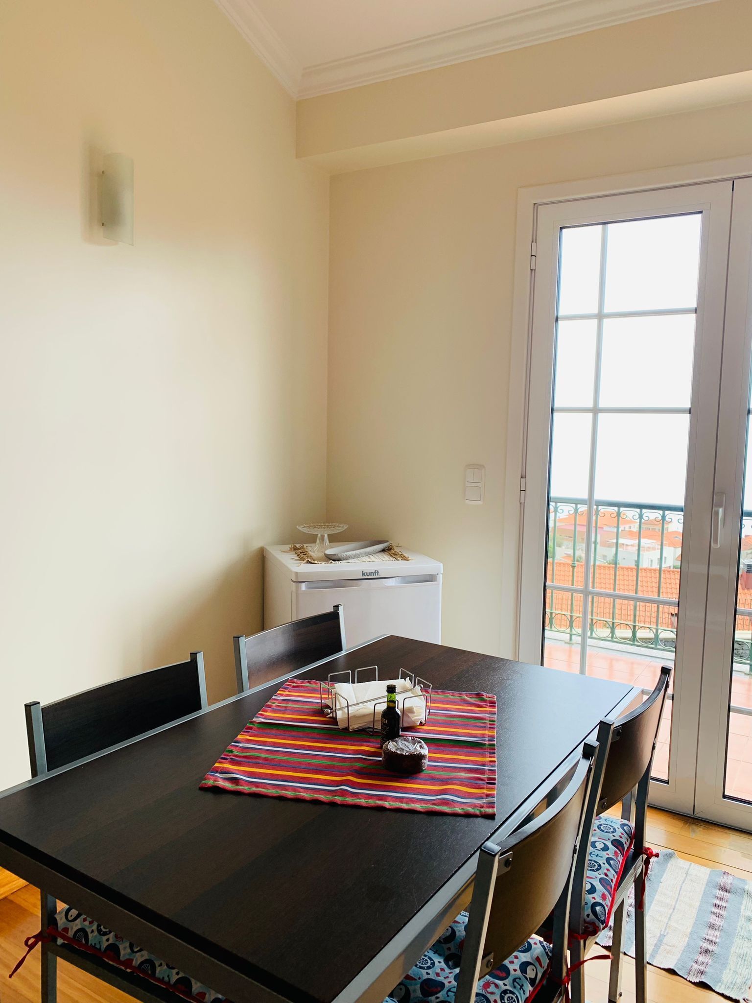 Un comedor con una mesa, sillas y un tapete colorido. Hay un electrodoméstico blanco pequeño en la esquina y vistas al balcón exterior.