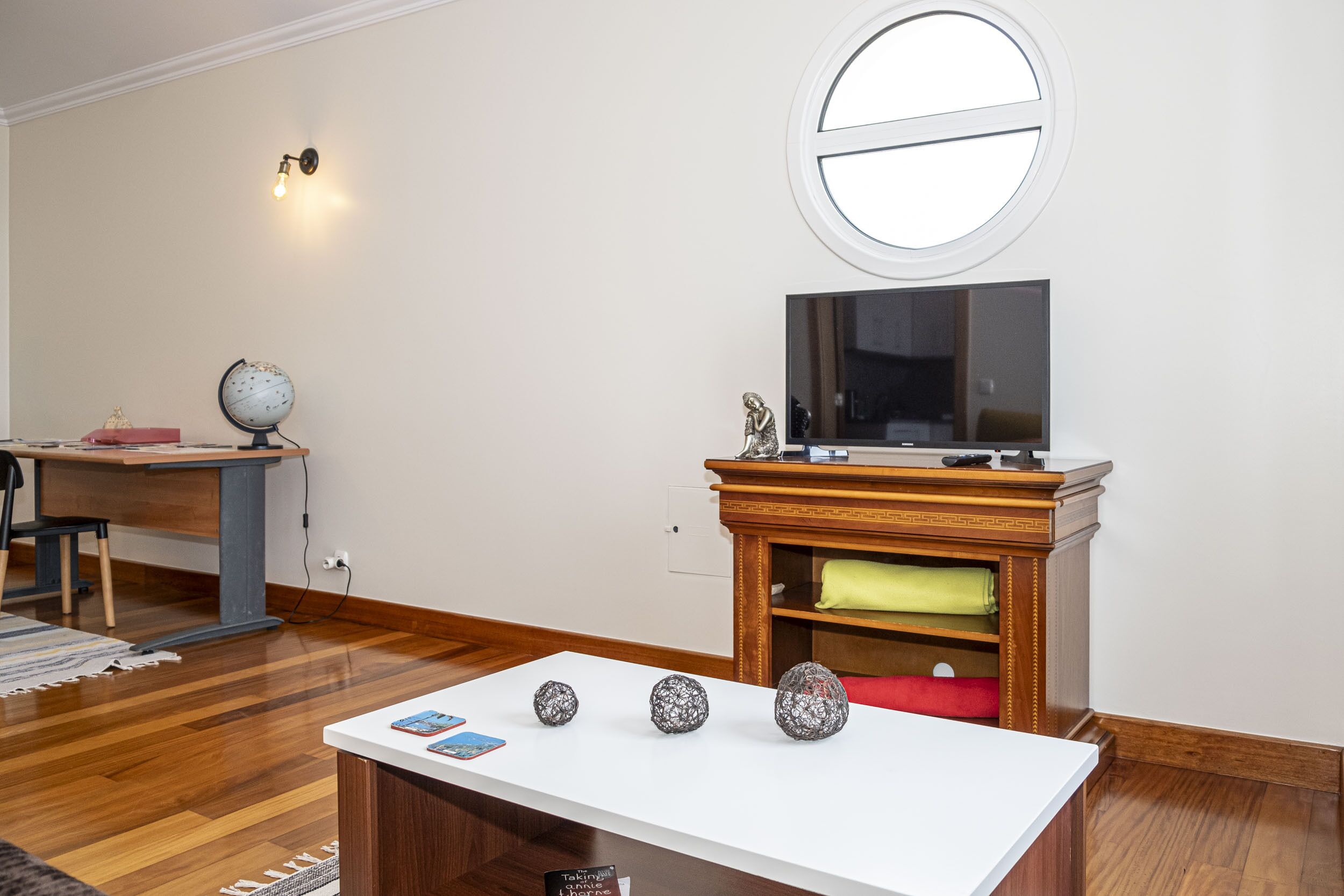 A living room with a white table displaying decorative items, a wooden TV stand with a flat-screen TV, a globe, and a statue on a desk.