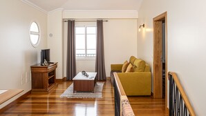A living room with a wooden floor, a sofa, a coffee table, a TV on a stand, and a window with curtains. - Harmony (Air conditioning, kitchen, balcony, self-check-in, dishware and linens provided, privacy) (Ponta do Sol)