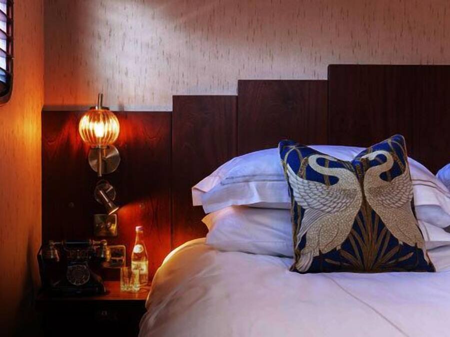 A hotel room with a wooden headboard, a nightstand with a lamp, and decorative pillows.