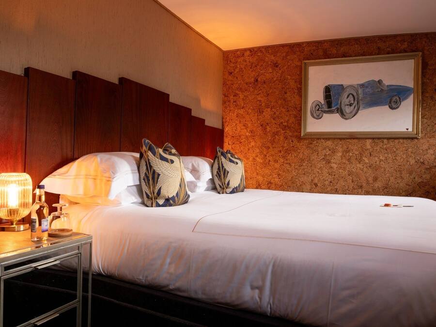 A hotel room with a bed, pillows, a nightstand with a lamp, a bottle, and a framed picture on the wall.