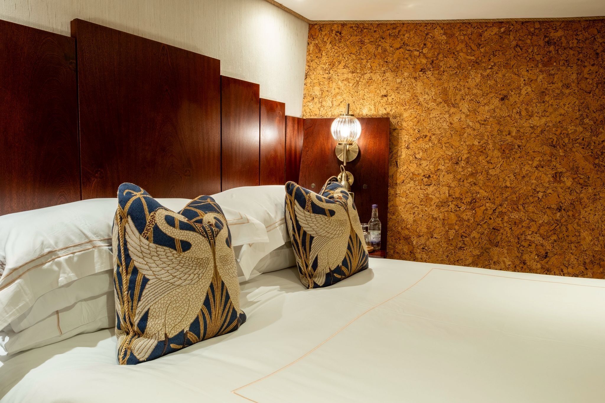 Una camera d'albergo con un letto grande, due cuscini decorativi, una testiera in legno e una parete con una texture simile al sughero.