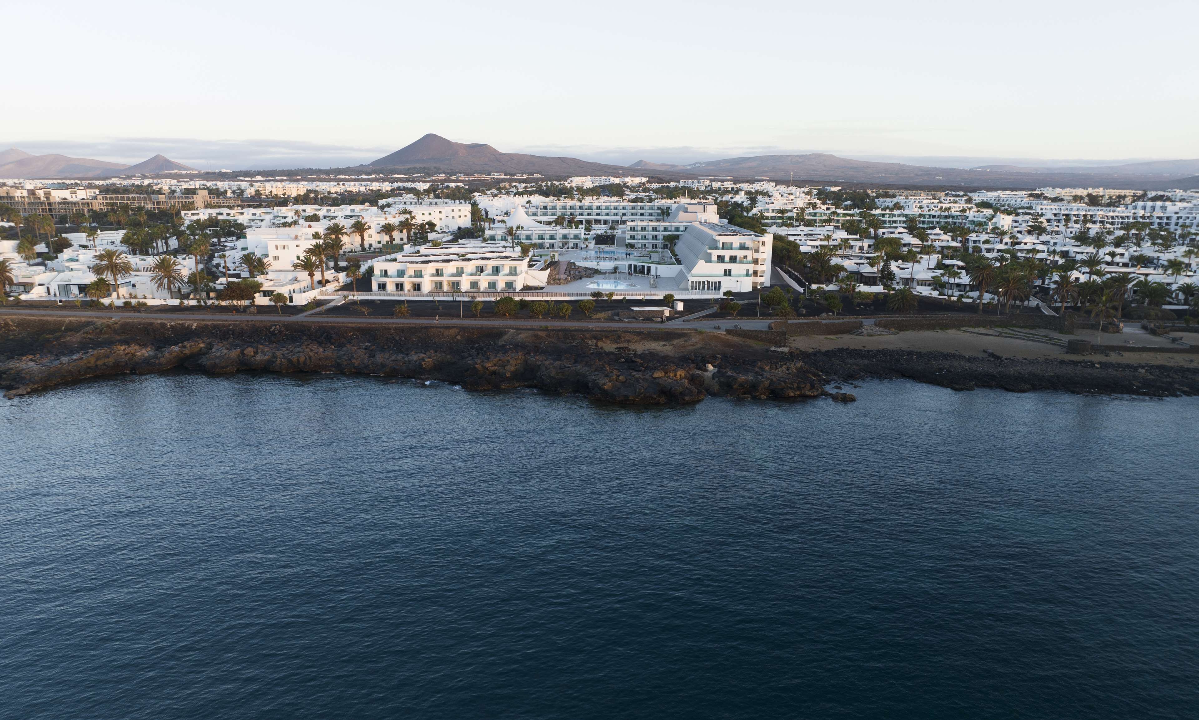 Vista aérea de un pueblo costero con edificios blancos, un hotel destacado con una estructura de techo singular, y una montaña al fondo.