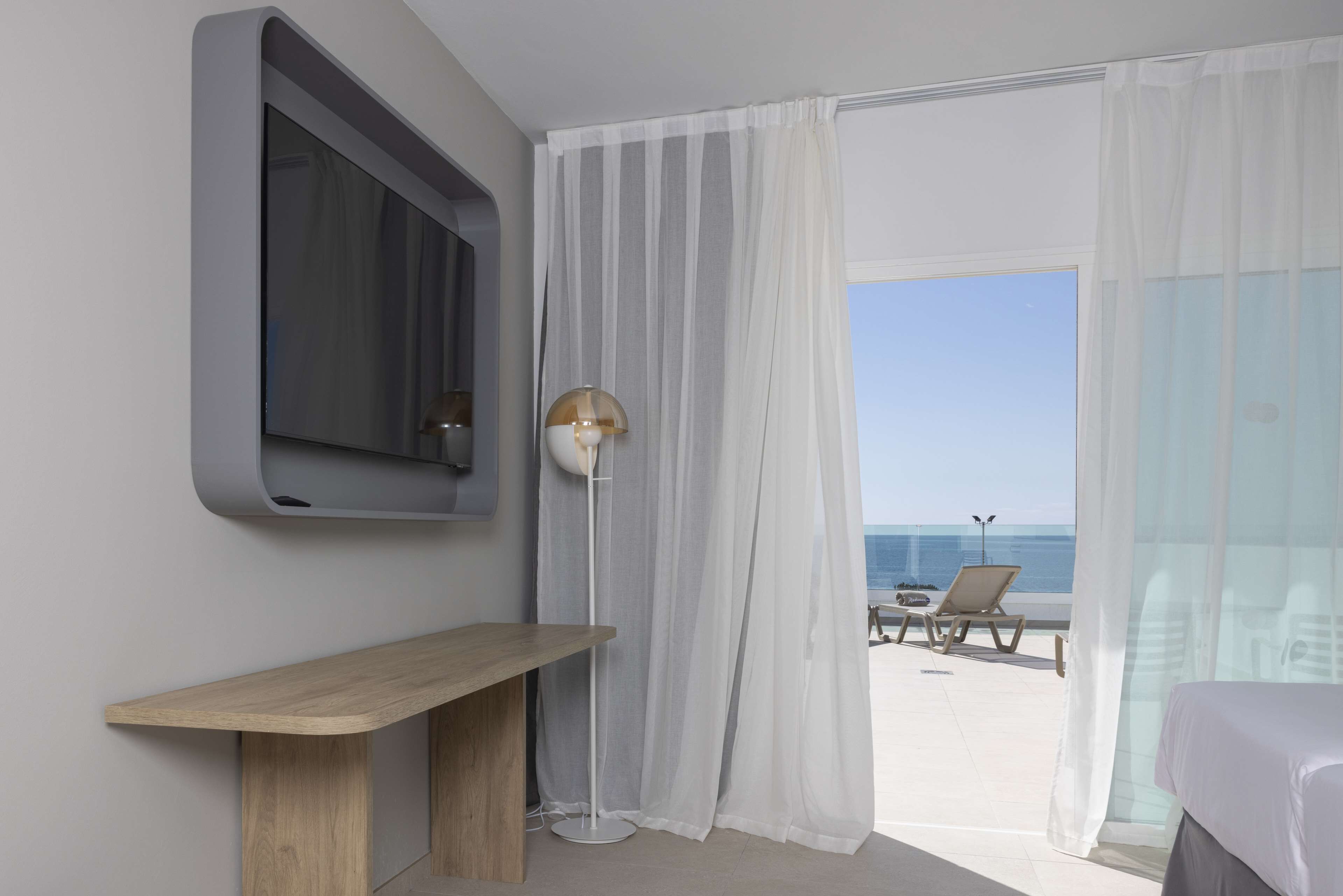 Habitación de hotel moderna con un televisor grande, una mesita de noche, una lámpara de pie y vista a la playa.