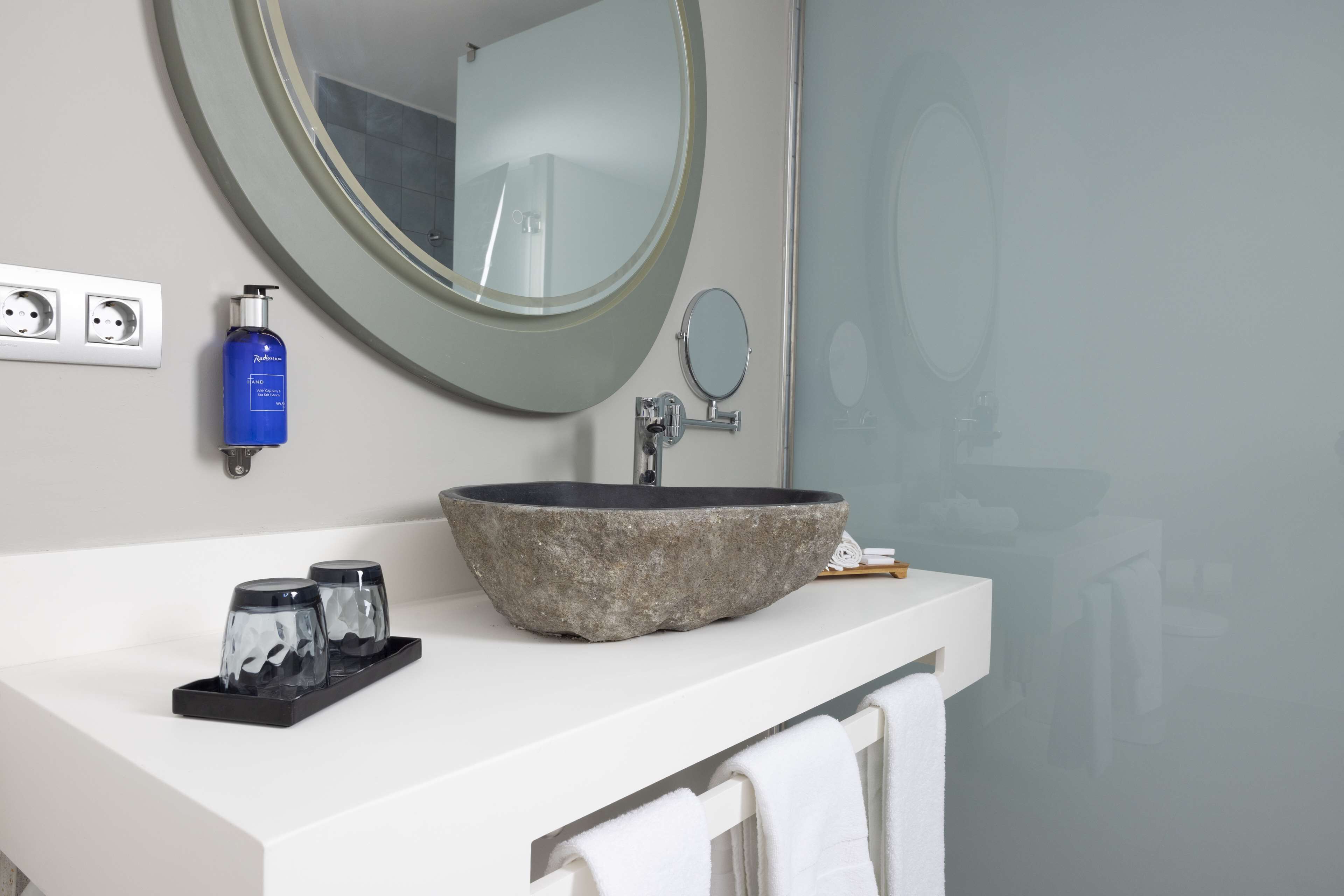A bathroom with a stone sink, a round mirror, a small round mirror, a bottle of hand soap, and a towel rack.