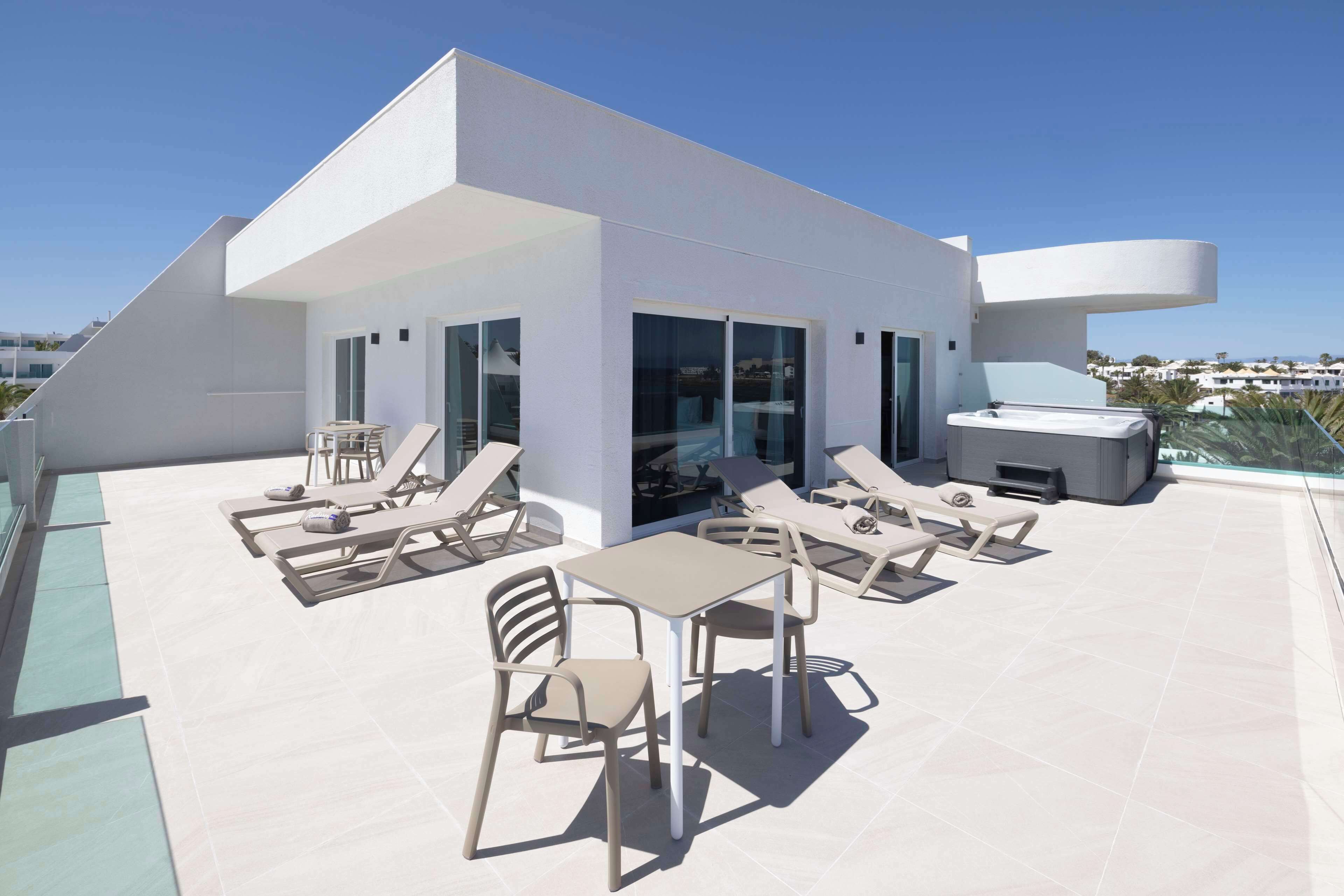 A modern rooftop terrace with sun loungers, a table, and a grill area.