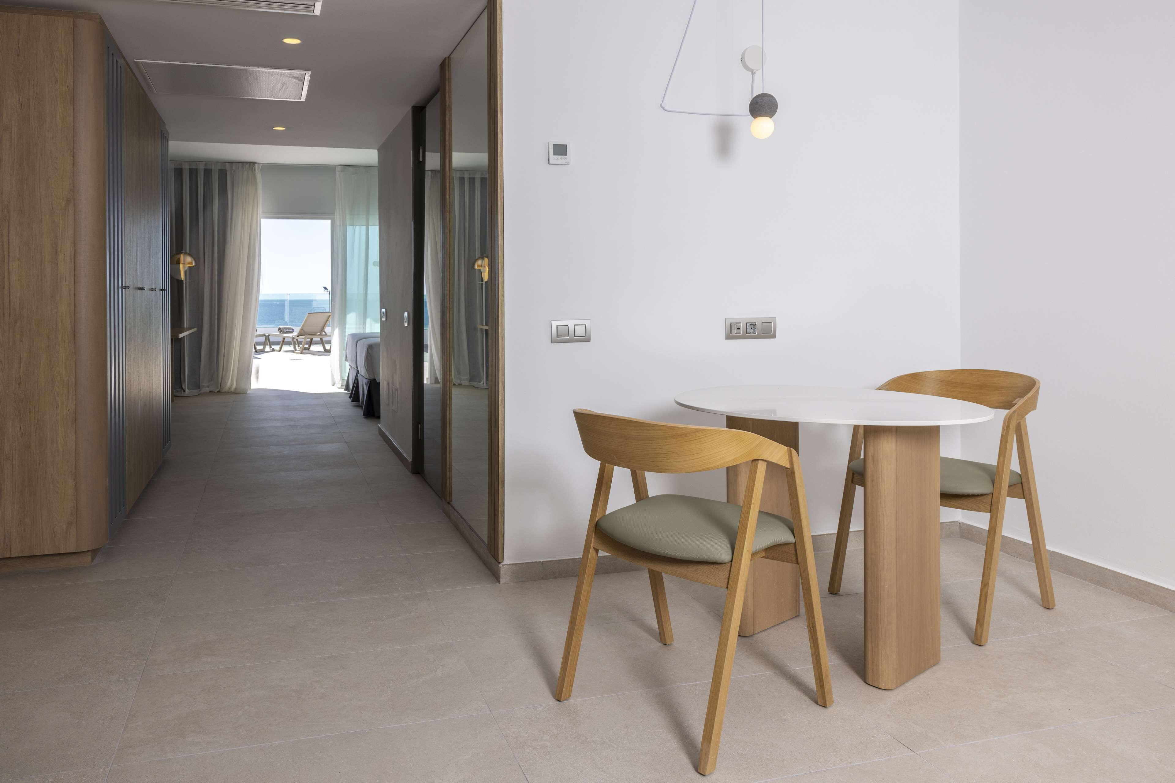 Una habitación de hotel moderna con un armario de madera, una mesa redonda de comedor con dos sillas y vistas al mar a través de ventanas amplias.