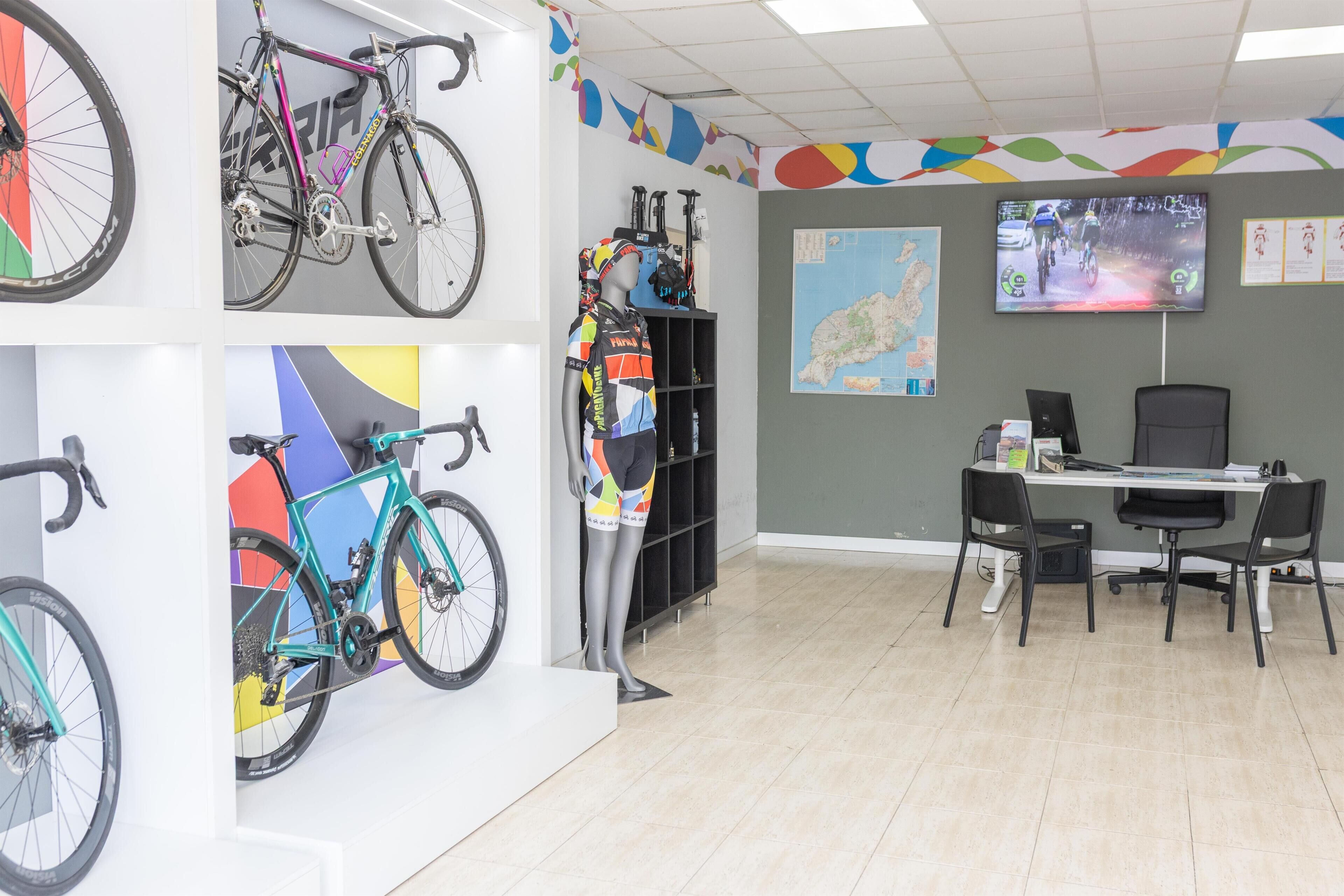 Ein Zimmer mit an der Wand befestigten Fahrrädern, einem Schreibtisch mit Stühlen und einem Fernseher, der eine Radveranstaltung zeigt.