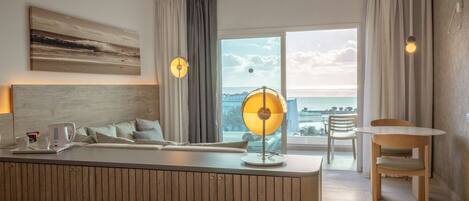 A modern hotel room with a wooden headboard, a large window with a view of the sea, a small dining area with a table and chairs, and a bedside lamp.