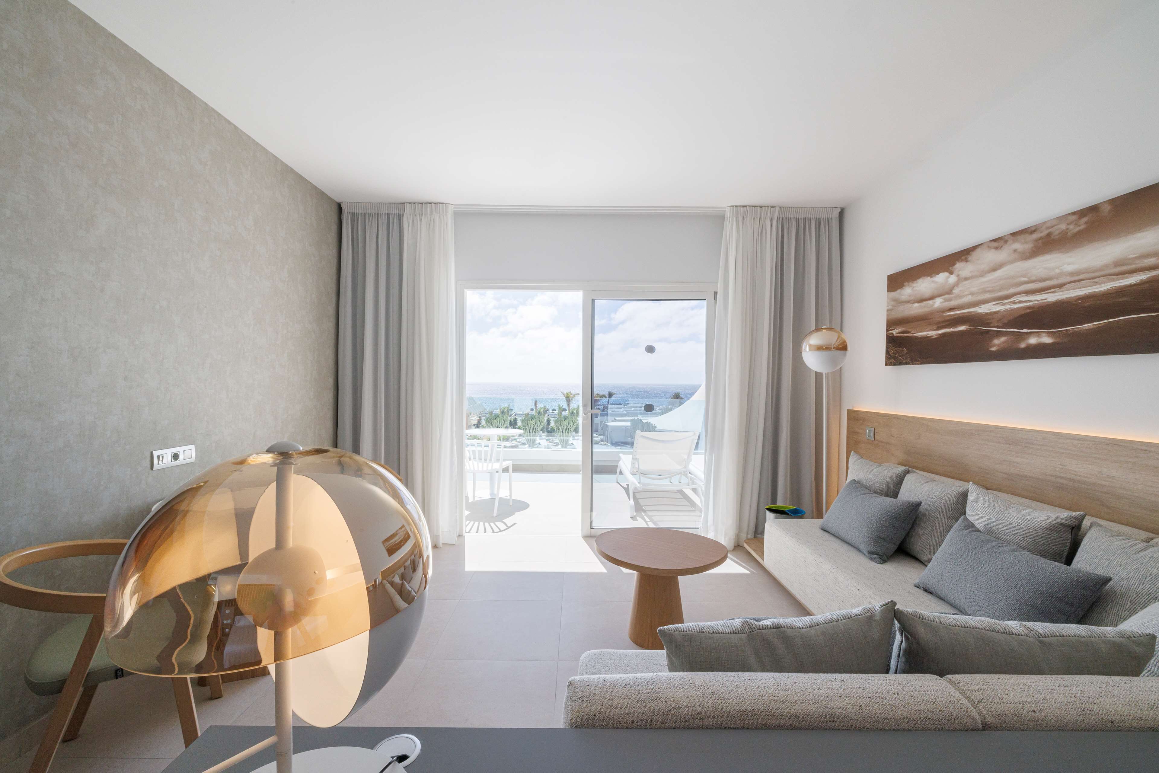 A modern hotel room with a balcony, a sofa, a round table, and a unique transparent lamp.
